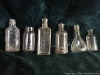 Six antique advertising glass bottles arranged horizontally on dark fabric, varying shapes and embossed markings visible.