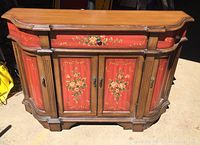 Front view of a wooden cabinet showing red painted panels decorated with floral artwork on drawer and double doors, wood frame with curved corners.