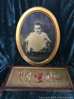 Full view of both items: oval baby photo with gold frame above rectangular motto in walnut frame below against dark blue cloth background.