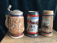Three ceramic steins on dark fabric background: Country Western Music with metal lid, 1991 Budweiser with winter scene, and original Gerzit West Germany with scenic artwork.