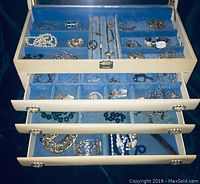 Photo of the open top section and drawers of a white jewelry box lined with blue velvet containing a variety of costume jewelry including necklaces, bracelets, rings, and a watch arranged in multiple compartments.