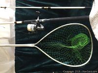 Fishing rod with foam grip and attached spinning reel lying on dark cloth background alongside large aluminum fishing net with green netting.