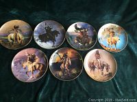 Seven decorative collector plates with Native American themed artwork by Hermon Adams, laid out on a dark green background.