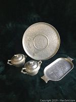 Four hammered aluminum items: one round tray with floral design, one rectangular tray with bamboo design, and a matched creamer and sugar bowl with lids and handles.