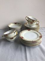 Four stacked plates, four stacked bowls, four stacked saucers, and four teacups arranged showing gold and burgundy leaf and scroll pattern trim