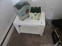 White side table with two decorative painted wooden pails on top, set against a wall and tan carpet.