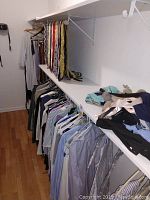 Closet rack with dress shirts hanging and folded pants and belts on white shelves