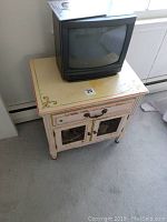 Photo showing tan and pink wooden side table with two doors and a drawer, measuring approximately 24x17x25 inches, with a Sharp CRT TV on top and visible wear on the furniture surface.