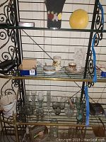 Black metal frame glass shelving unit with an assortment of glassware on three shelves including various candle holders, glass cups, vases, and ceramic ware.