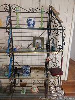 Metal black shelving unit holding multiple items including Asian blue and white ceramic planter, candle holders, lockable box, pig frame, bowls, glasses and decorative items