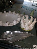 Photo showing ceramic Easter bunny bowl with multiple bunnies sculpted on outer edge, white ceramic fruit bowl, wooden green bird sculpture, and silver-toned metal leaf tray.
