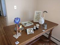 Wide view showing all assorted home decor items on wooden table: colorful abstract floral clock, pewter cup, ceramic pitcher, candle holders, metal grape dish, decorative duck tray, silver desk clock, and a white framed wall clock.