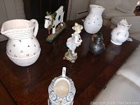 View of all items on a dark wood table including two white pitchers with blue dots, a small gold pitcher, a porcelain girl figurine, a wooden cow decor piece, and a small blue and white pitcher.