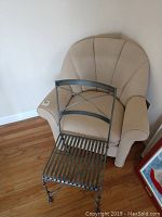 Photo showing a beige upholstered armchair behind a black metal chair on a hardwood floor near the wall.