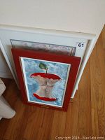 Photo showing three framed artworks leaning against a white piece of furniture and chair. The front artwork is 'Adams Apple' featuring a red apple core with human-like breast imagery in a reddish wooden frame with a gold trim.