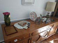 Wide view of the wooden sideboard with various decorative items arranged on top: October Hill floral vase with pink roses, wooden mesh tray, decorative plate, candle holders, a metal tissue box, a small glass bowl mirror, and a 26-inch lamp with beige shade