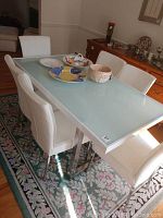 Photo showing the dining table with frosted glass top and metal frame surrounded by six white leather chairs placed on a patterned area rug.