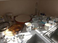 Overview of assorted mugs, glass and ceramic kitchenware, cow napkin holders, jars, salt and pepper shakers, and metal paper towel holder arranged on a counter under a window with daylight.