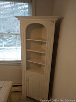 Full view of the white corner cabinet with four shelves, showing its overall size and design by window.