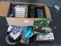 Box of assorted household and small electronics items including Coleman lantern, small TV, coin counter, plates, and kitchen towels.
