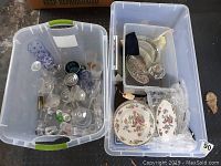 Two large plastic bins, one contains assorted glassware items including various glasses and crystal pieces, the other contains china with floral patterned plates and clear glass serving dishes.