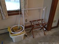 View of all main items including wooden luggage rack, white plastic clothes rack on wheels, stacked laundry and waste baskets, and white curtain rods leaning on the wall.