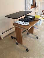 Photo showing rolling computer table with wood grain finish holding three surge protectors and cords, and partially visible adjustable black bed table above
