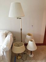 Four lamps in group shot: tall brass floor lamp with white pleated shade and swivel arm; three table lamps with white or beige shades and brass or decorative bases.