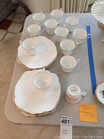 Top-down view of 12 white fine bone china plates with scalloped edges and gold trim, along with matching 12 teacups with gold trim arranged on top.