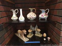 Photo showing the three china pitchers, two vases, onyx jaguar figurine, onyx bookends, and small decorative mushroom items arranged on a reflective glass surface.