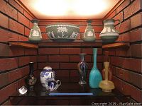 Shelf display of green Wedgewood jasperware pieces and assorted glass vases on two shelves against red brick wall.