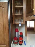 Photo showing four vintage thermos bottles on kitchen counter under opened cabinet with drinking glasses inside the cabinet.