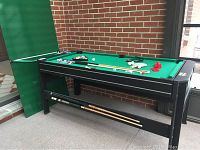 Black multi-game felt table with green pool playing surface with cues, pool balls, ping pong paddles, and air hockey pucks arranged on top and below the table.
