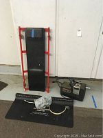 Overview of the lot showing the red-framed auto creeper, black air compressor, black plastic battery carrier, and metal grease gun on black mat.