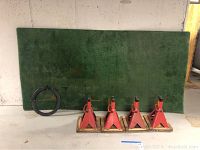 Green usage-worn astro turf panel leaning against wall with a black circular drain snake and four red Super Lift automotive jack stands on wooden bases lined up on floor.