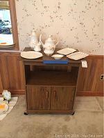 Full view of china set on wooden rolling cart with two doors and shelf.