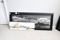 Two cityscape prints leaning against a wall, one large black and white with a tower and skyline, the other smaller with gold bridge accents.