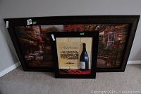 Two framed prints leaning against a wall, one larger with a vineyard outdoor cafe scene and one smaller with a wine bottle and text.