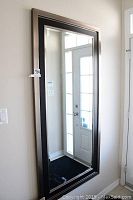 Large rectangular metal framed wall mirror mounted on a white wall in an entryway, reflecting front door and window light.