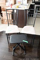 Two black metal frame chairs with light beige padded seats alongside one black adjustable stool with padded seat and five-star wheeled base shown on hard floor.