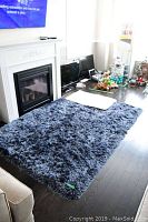 Blue shag carpet rug laid on dark wooden floor in front of fireplace and couch, with household items in background.