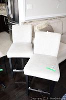 Two beige fabric bar stools placed side by side in front of a beige fabric sofa with decorative pillows. Dark hardwood flooring is visible, showing the stools' black wooden legs and metal footrests.