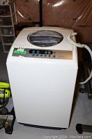 Front view of white Midea top-load washer with glass lid, control panel, and attached hoses on the side.