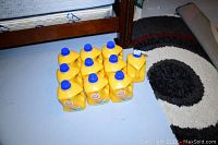 Ten yellow plastic jugs of Arm and Hammer liquid detergent with blue caps arranged in two rows on a floor next to furniture and a rug.