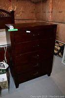 Wooden five drawer dresser shown from the front with drawers closed in a dimly lit basement room.