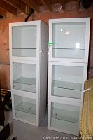Two closed white shelving units side by side against unfinished wall, each with three glass shelves visible through framed doors.