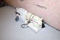 Two sewing machines side by side on floor against wall showing cords and pedal for each machine