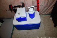 View of large blue and white cooler with smaller lunchbox cooler and water jug on top, metal cart behind.