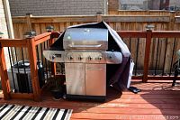 Front and side angled view of a stainless steel BBQ grill with four main burners, side burner, double-door storage, and a protective cover partially draped over it, placed on wooden deck.