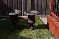 Two rusted metal urns displayed outdoors on grass next to a wooden fence and red wood decking. Both urns have classical design with flared rims and square bases.